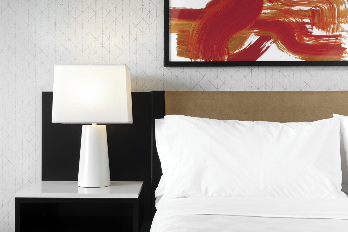 A neatly made bed with white linens, a bedside table with a white lamp, and a framed abstract painting on the wall.