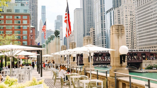 An outdoor dining area with tables and umbrellas, overlooking a river with a bridge and high-rise buildings in the background. - Holiday Inn Chicago Dwtn - The Mart by IHG (Chicago)