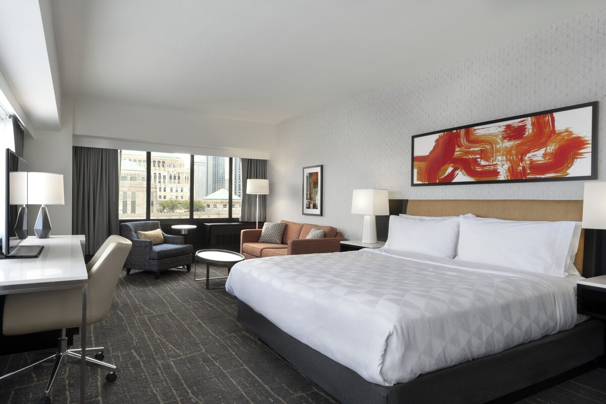 A hotel room with a large bed, a desk, a chair, a sofa, and a window with a city view.