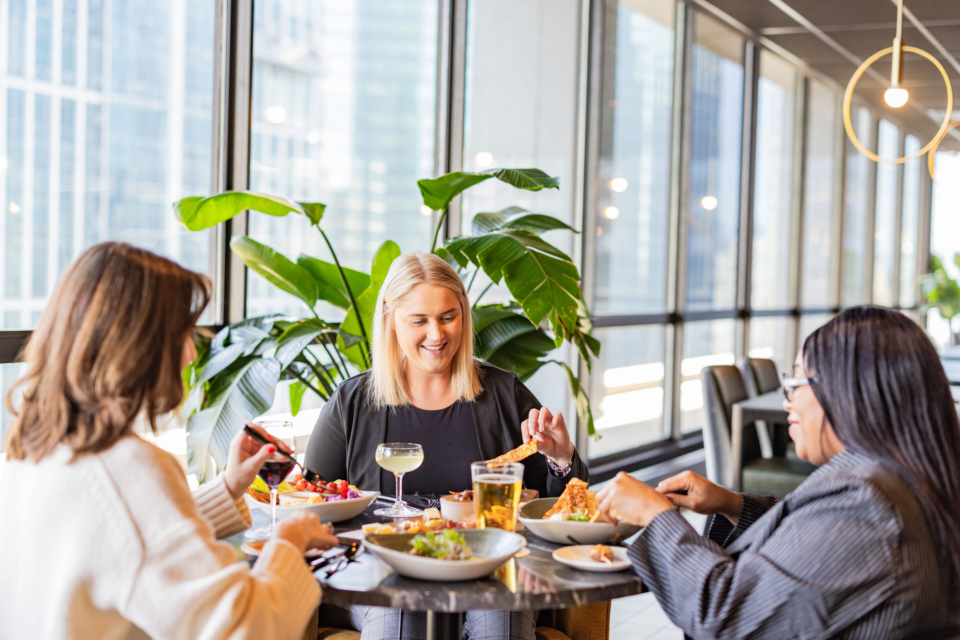 Un grupo de personas cenando en una mesa con diversos platos, bebidas y un amplio ventanal con vistas a edificios.