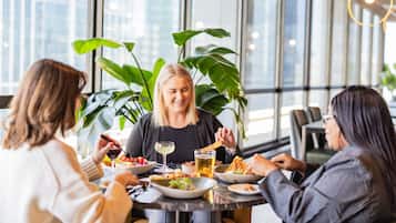Un grupo de personas cenando en una mesa con diversos platos, bebidas y un amplio ventanal con vistas a edificios.