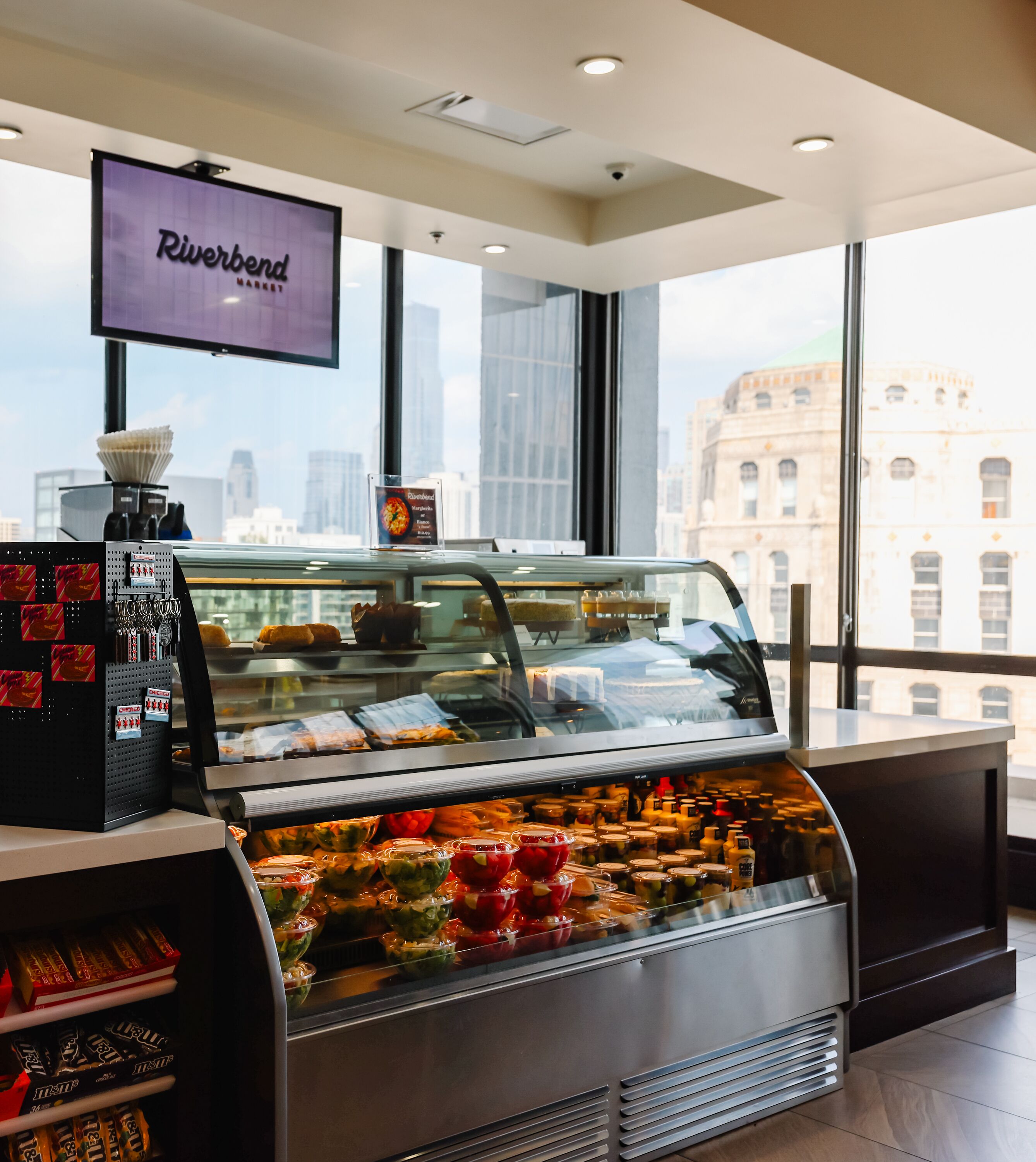 A Riverbend Market display case with various food items and condiments, a television above it, and a city view through large windows.