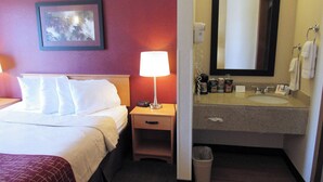 A hotel room with a bed, bedside lamp, nightstand, and a bathroom with a sink and mirror.