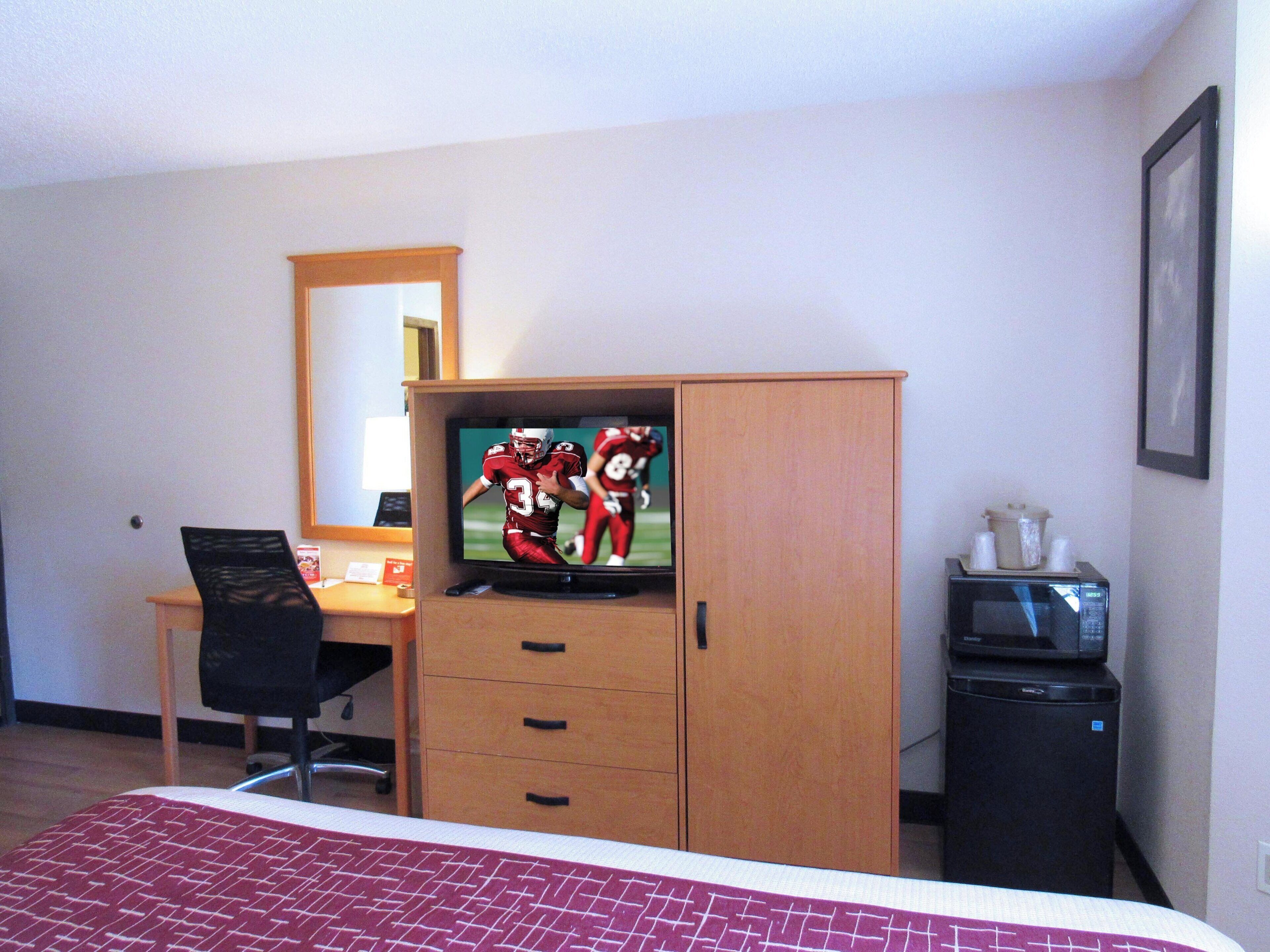 Habitación de hotel con cama, escritorio con computadora, televisor, espejo, microondas y refrigerador.
