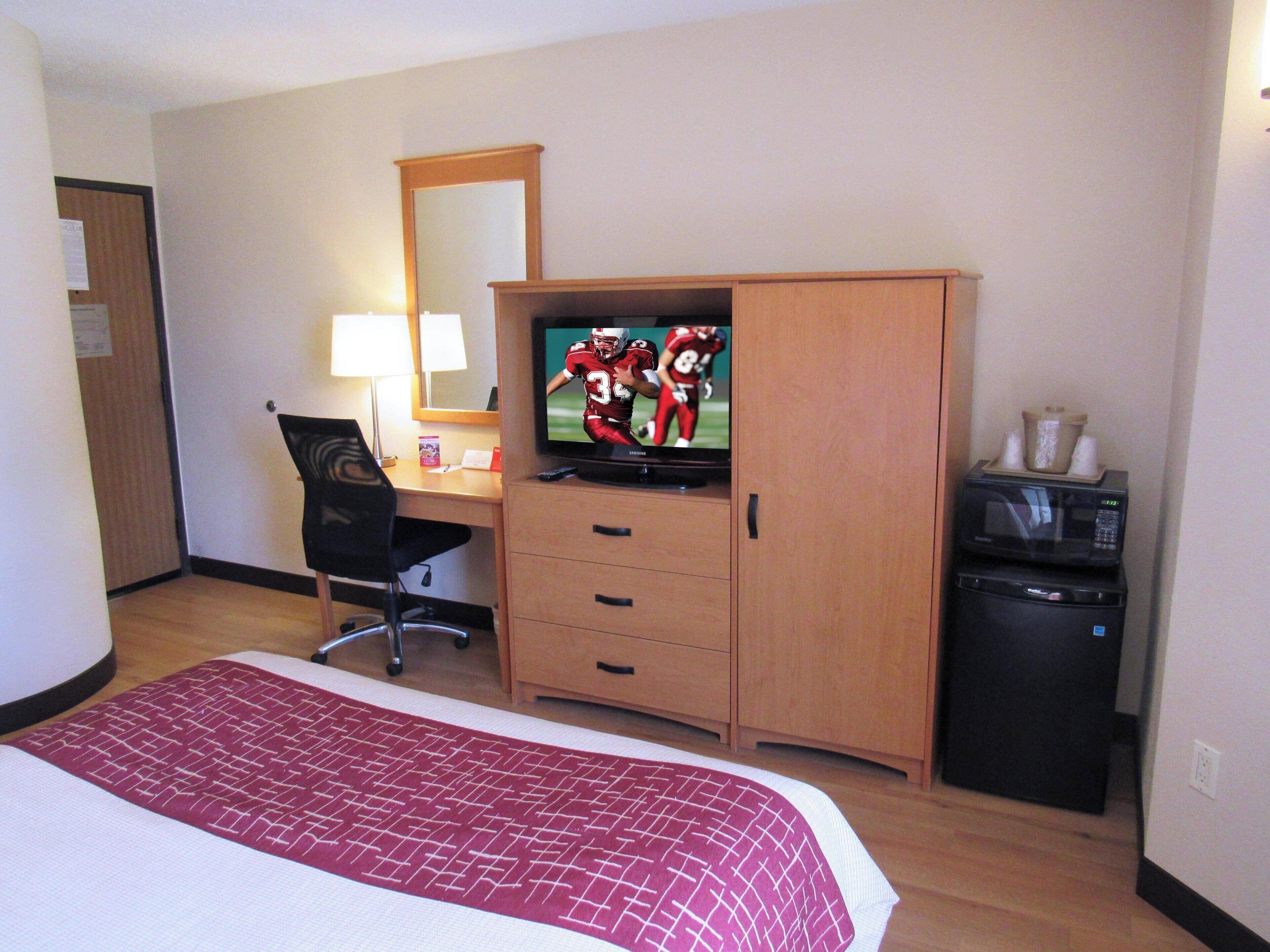 A hotel room with a bed, a desk with a chair, a television, a microwave, and a small trash bin.