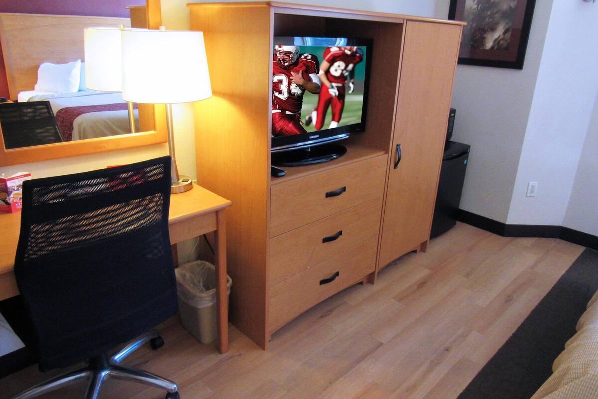 A hotel room with a wooden desk, a chair, a television on a cabinet, a lamp, and a trash can.