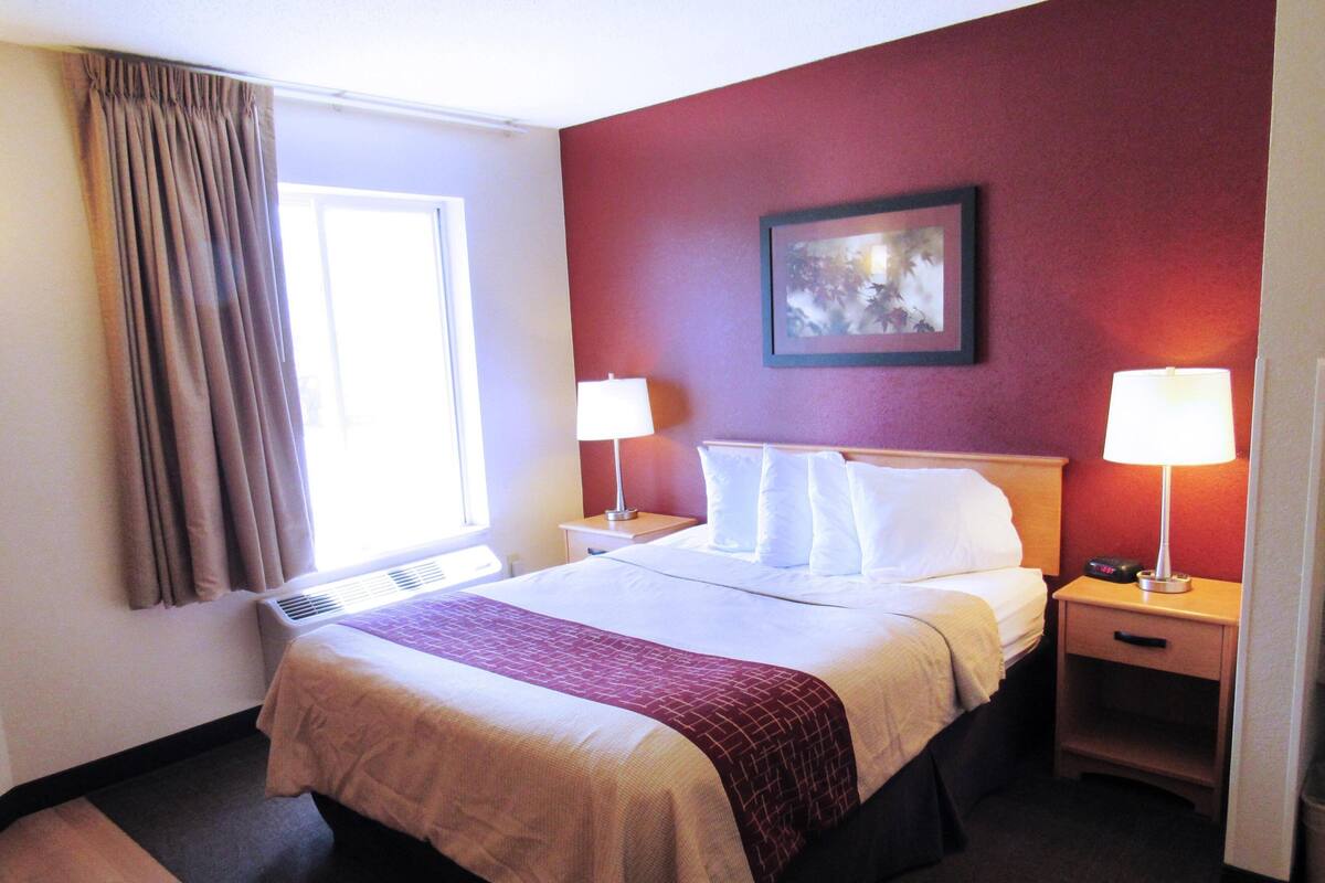 A hotel room with a bed, two bedside tables with lamps, a window with curtains, a wall-mounted picture, and a carpeted floor.