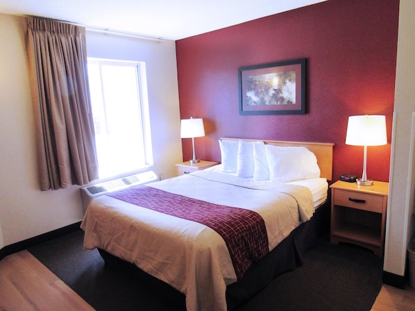 A hotel room with a bed, two bedside tables with lamps, a window with curtains, a wall-mounted picture, and a carpeted floor.