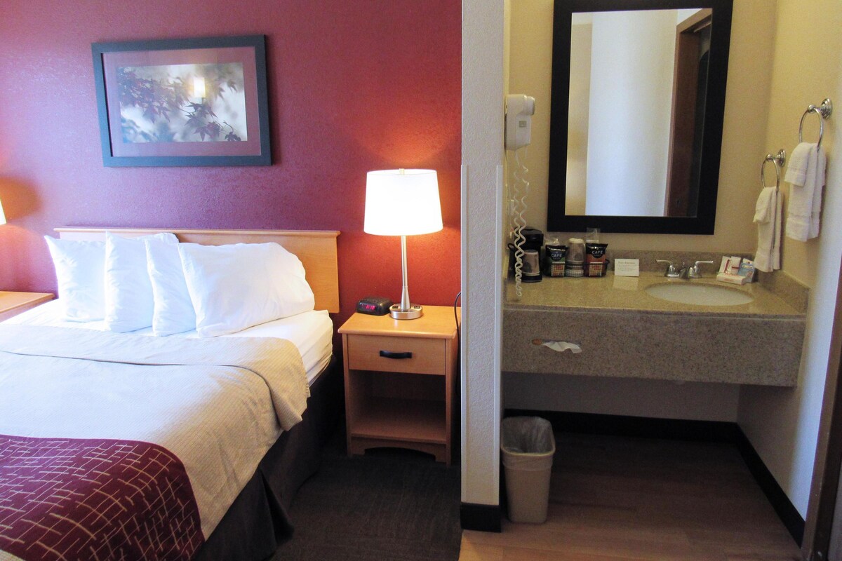 A hotel room with a bed, bedside lamp, nightstand, and a bathroom with a sink, mirror, and towel rack.