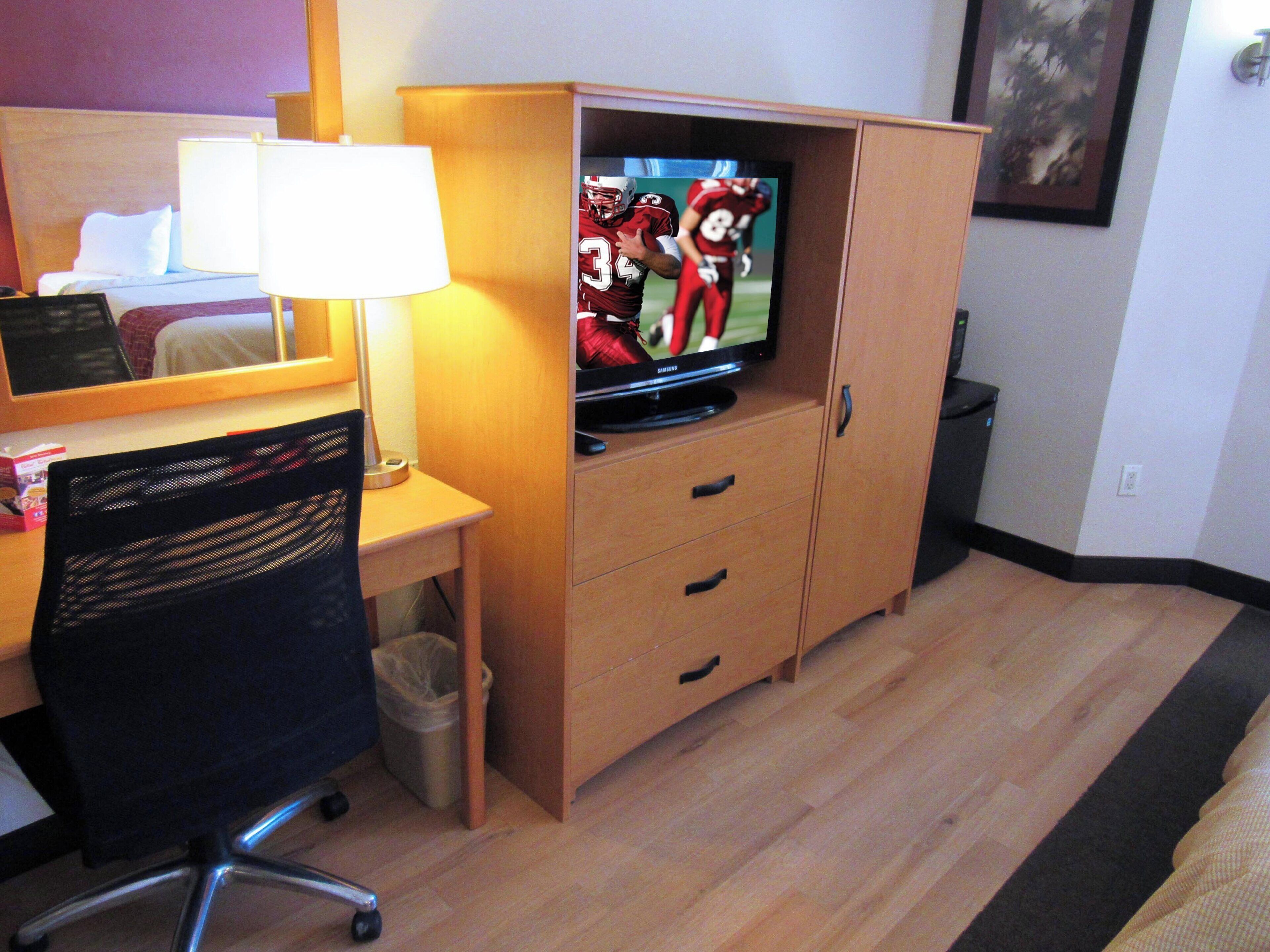A hotel room with a wooden desk, a chair, a television on a cabinet, and a bed with a white pillow.