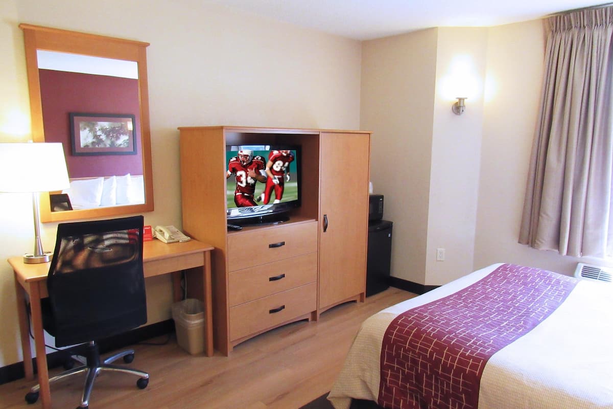 A hotel room with a wooden desk, a television, a bed with a patterned blanket, and a window with curtains.
