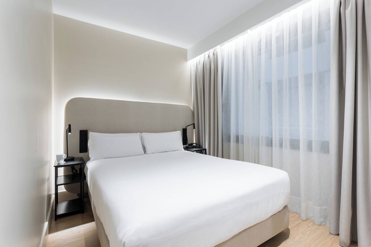 A modern hotel room with a large bed, a bedside table with a lamp, and sheer curtains.