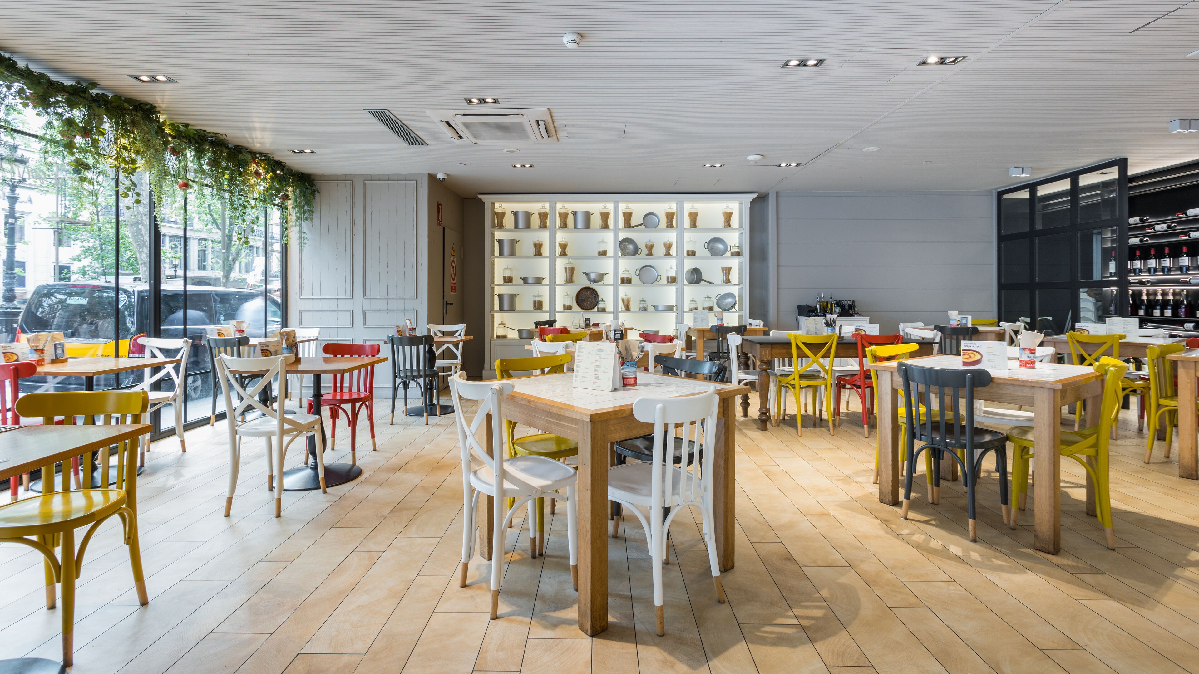 Un ristorante con pavimento in legno, sedie bianche e colorate e una parete che espone utensili da cucina.