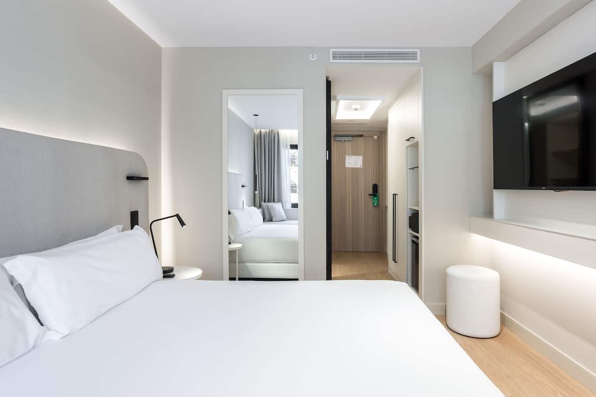 A modern hotel room with a large bed, bedside table, lamp, and a flat-screen TV mounted on the wall.