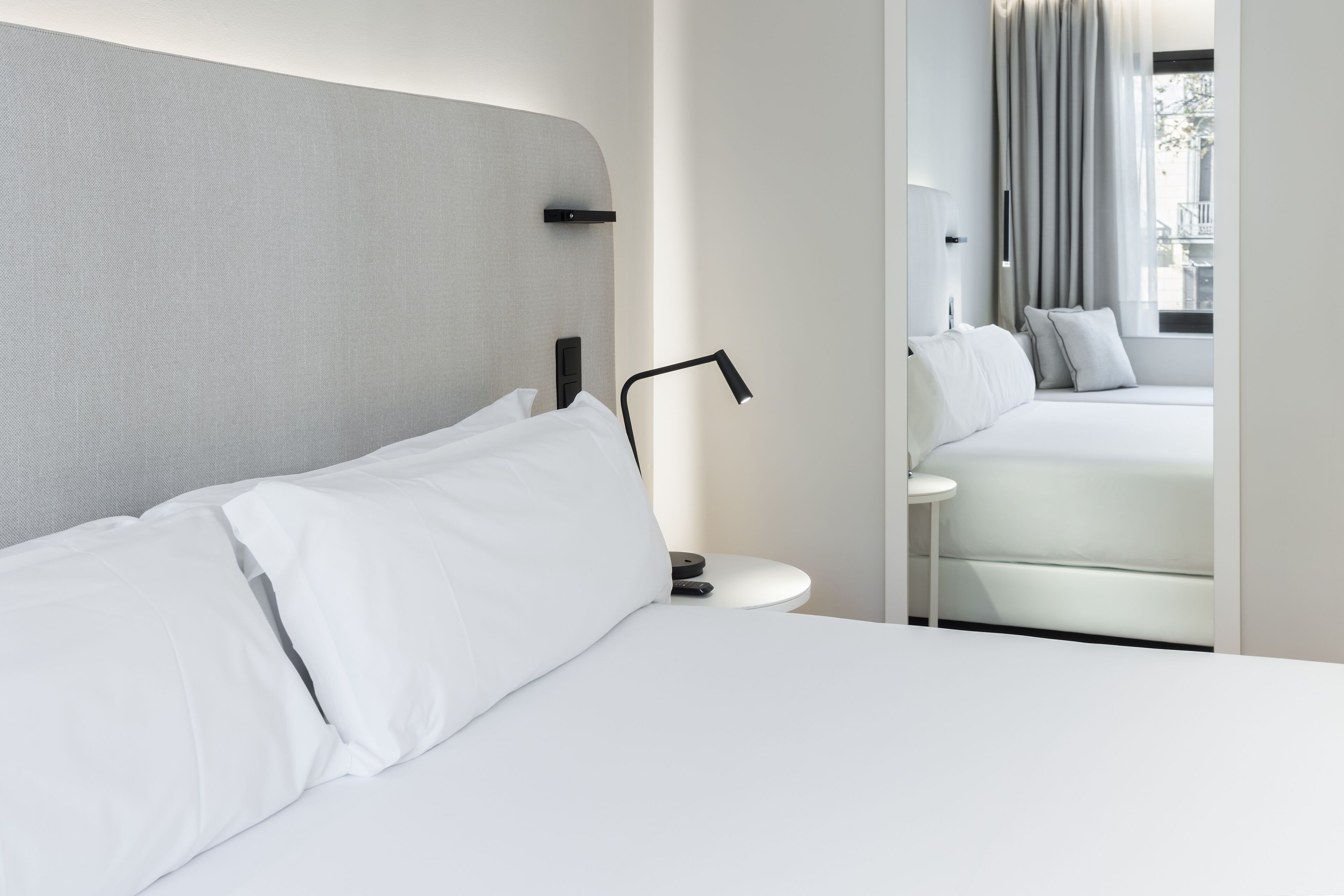 A modern hotel room with a neatly made bed, a bedside table with a lamp, and a view of a balcony with curtains.