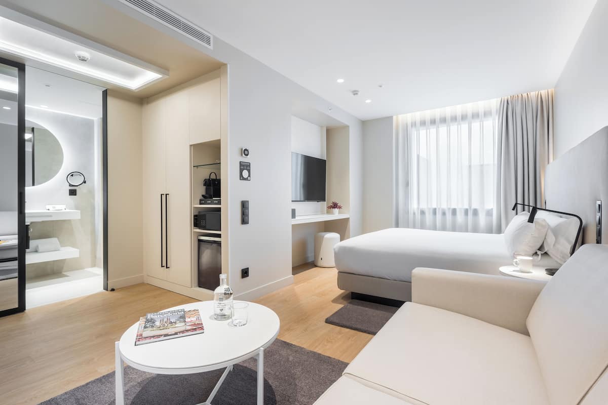 A modern hotel room with a bed, a small table with a magazine, a TV, and a bathroom with a mirror and toilet.