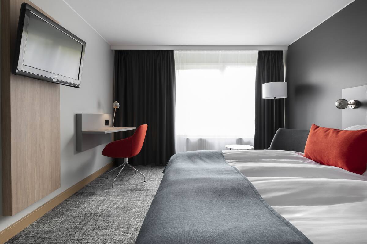 A hotel room with a bed, a desk, a chair, a TV, and a window with curtains.