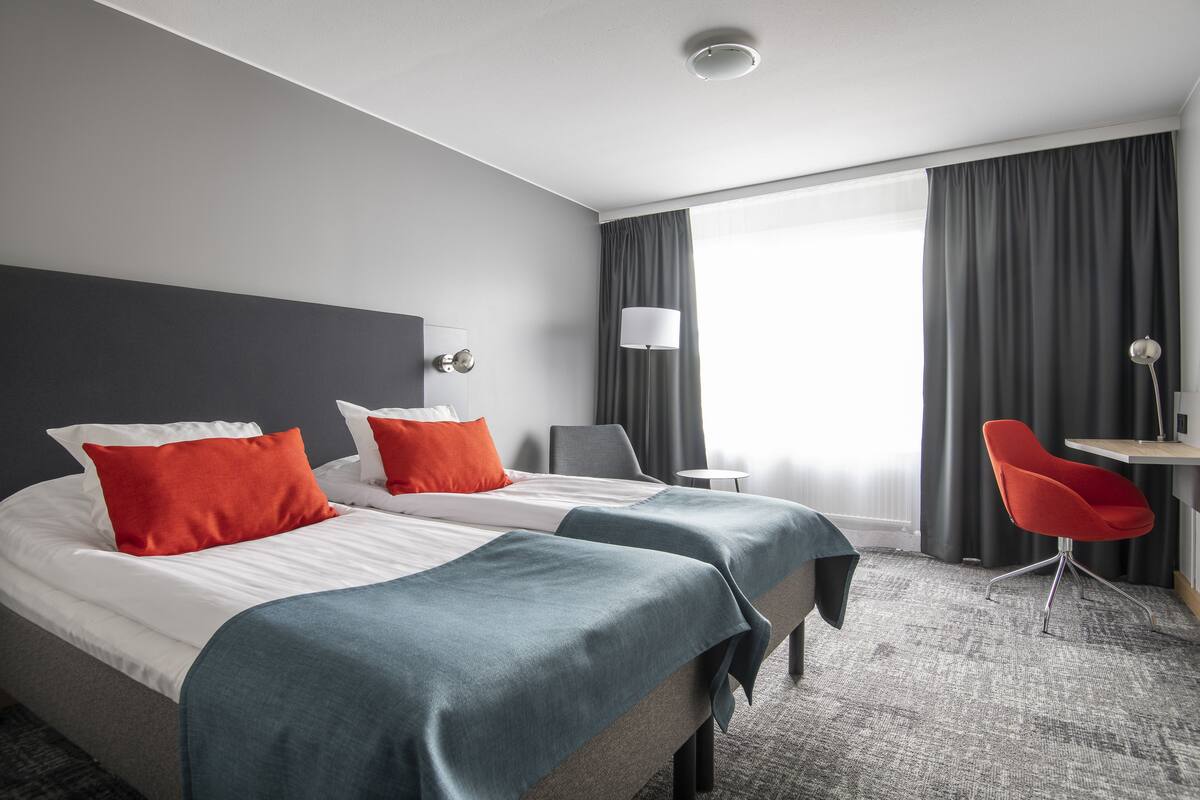 A hotel room with two beds, a desk, a chair, a lamp, and a large window with curtains.