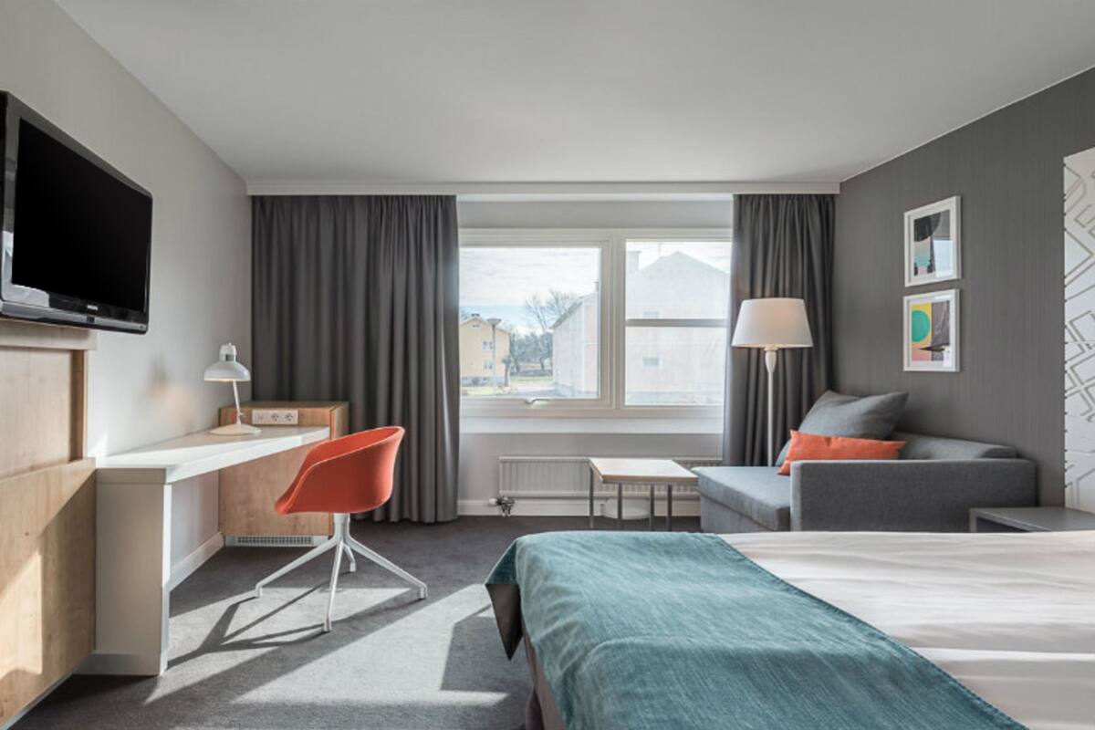 A hotel room with a bed, a desk, a chair, a TV, a lamp, and a window with curtains.