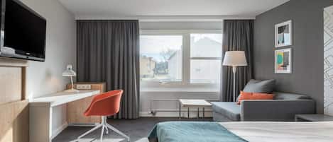 Habitación de hotel con cama, escritorio, silla, televisor, lámpara y ventana con cortinas.