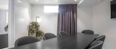 A conference room with a long table, six chairs, a potted plant, a window with curtains, and a mounted TV.