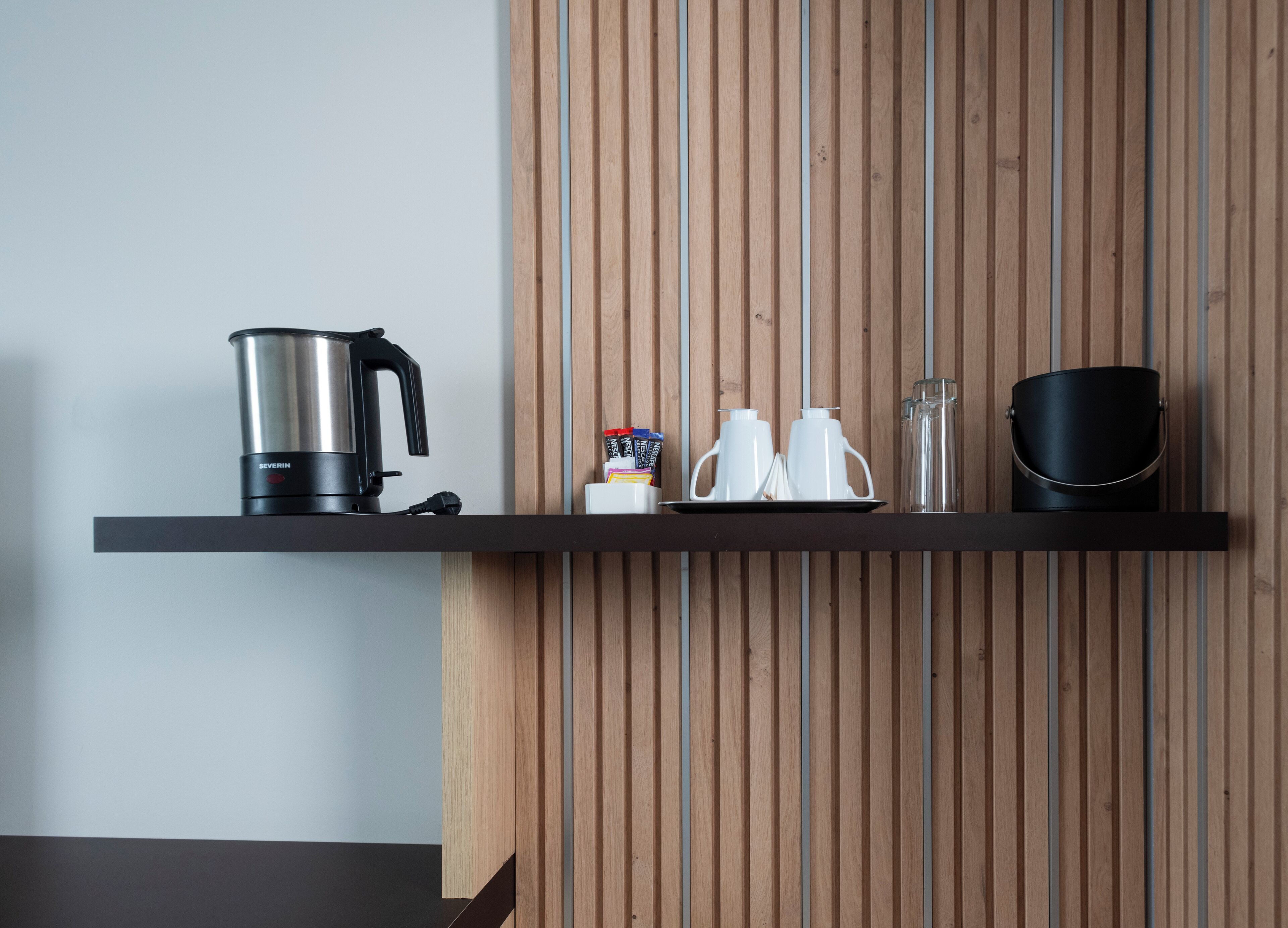 A kitchen shelf with a coffee maker, two white teapots, a glass, and a black container.
