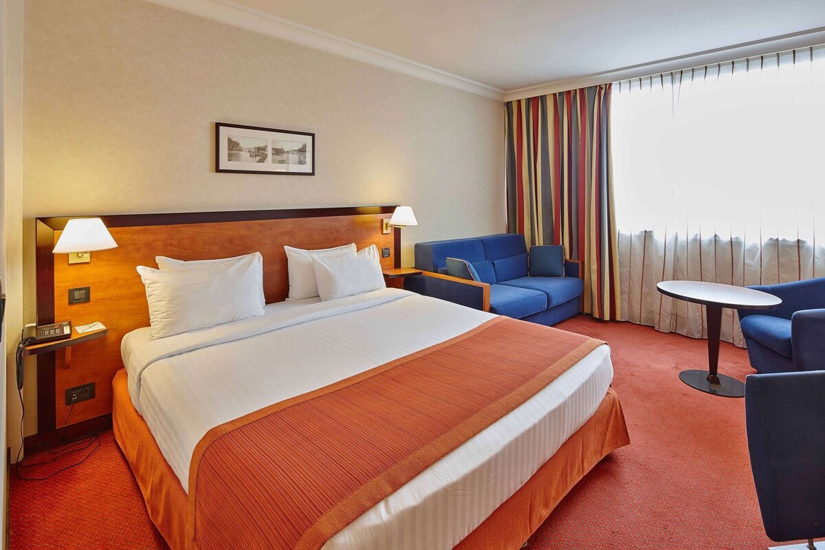 Una habitación de hotel con una cama grande, dos lámparas de noche, un sofá azul, una mesita redonda y una ventana con cortinas a rayas.
