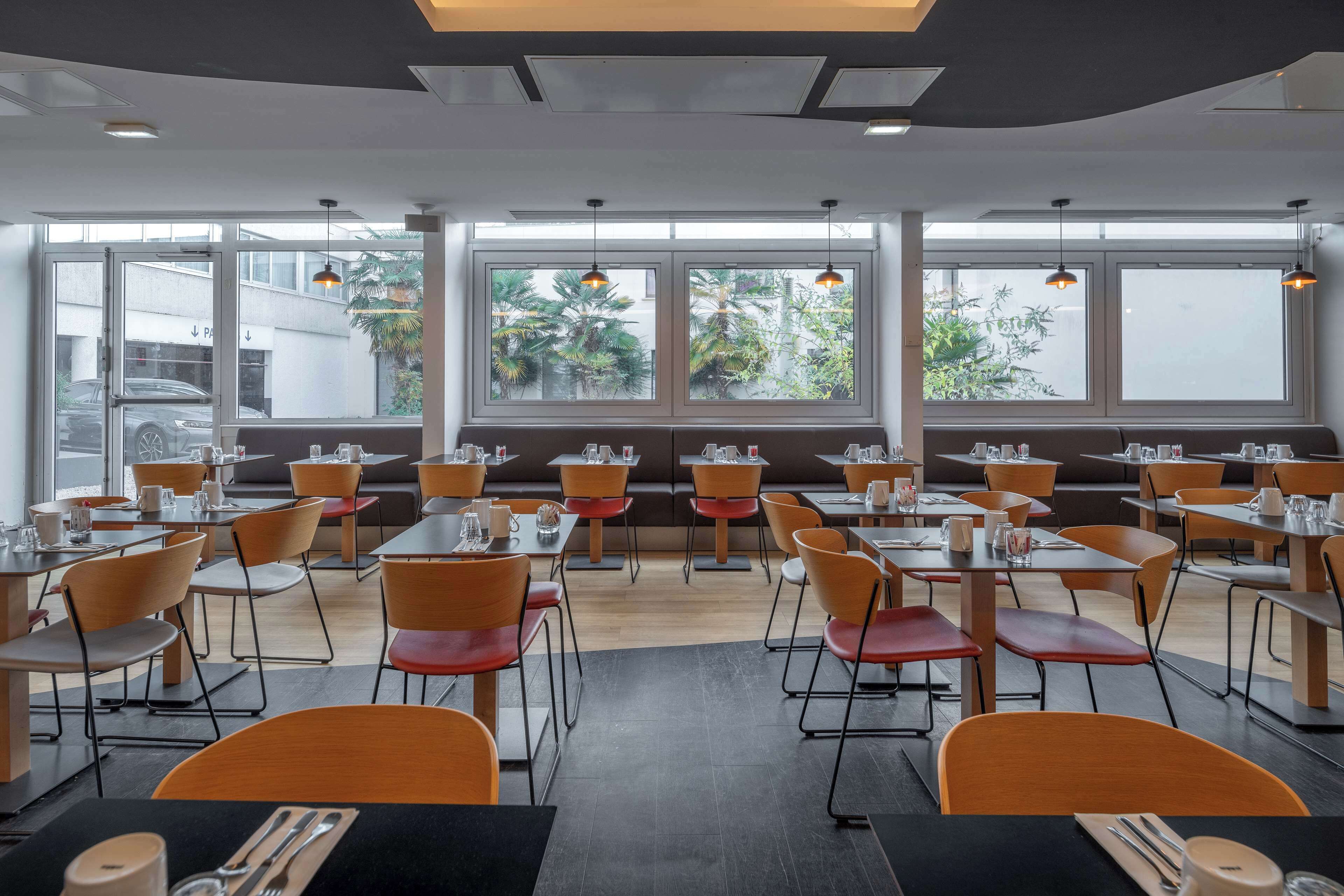 A modern dining area with multiple tables and chairs, large windows with views of greenery, and pendant lighting.