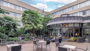 Ein Terrassenbereich im Freien mit Tischen und Stühlen, umgeben von Grünpflanzen und einem Gebäude mit großen Fenstern.