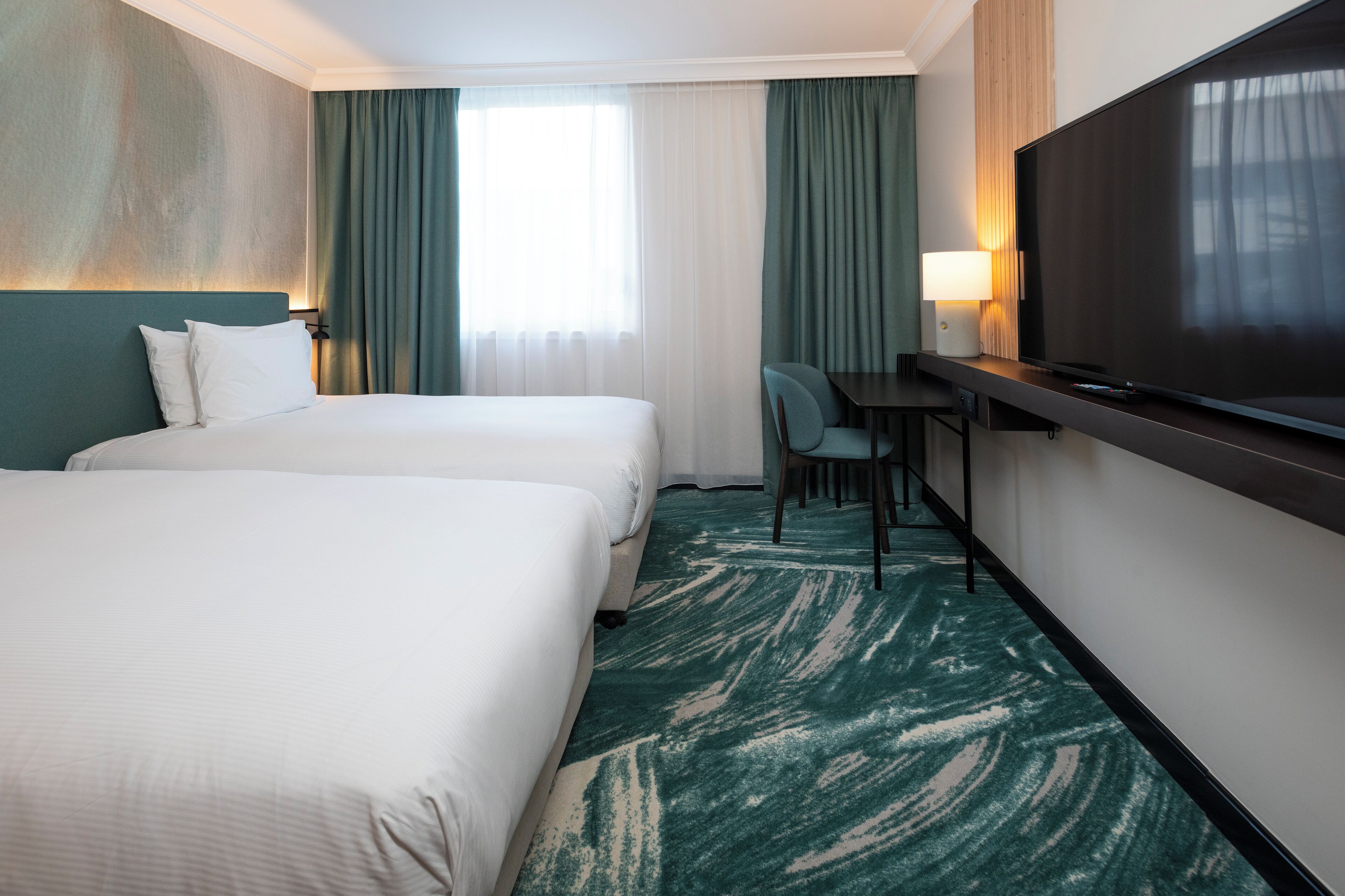 Habitación de hotel con dos camas, un escritorio con silla, un televisor y un ventanal con cortinas.