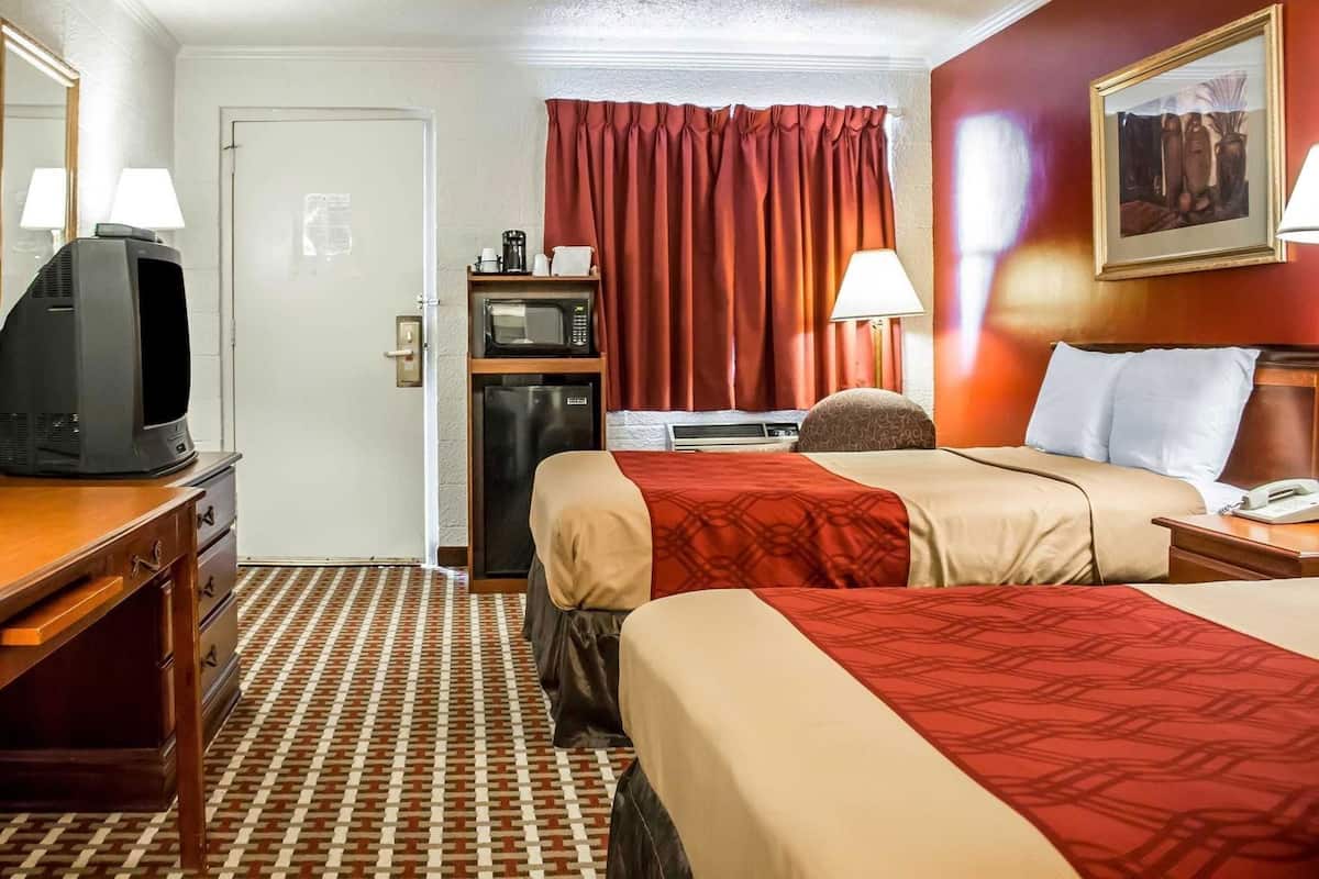 A hotel room with two beds, a desk with a television, a refrigerator, a microwave, a lamp, and a painting on the wall.