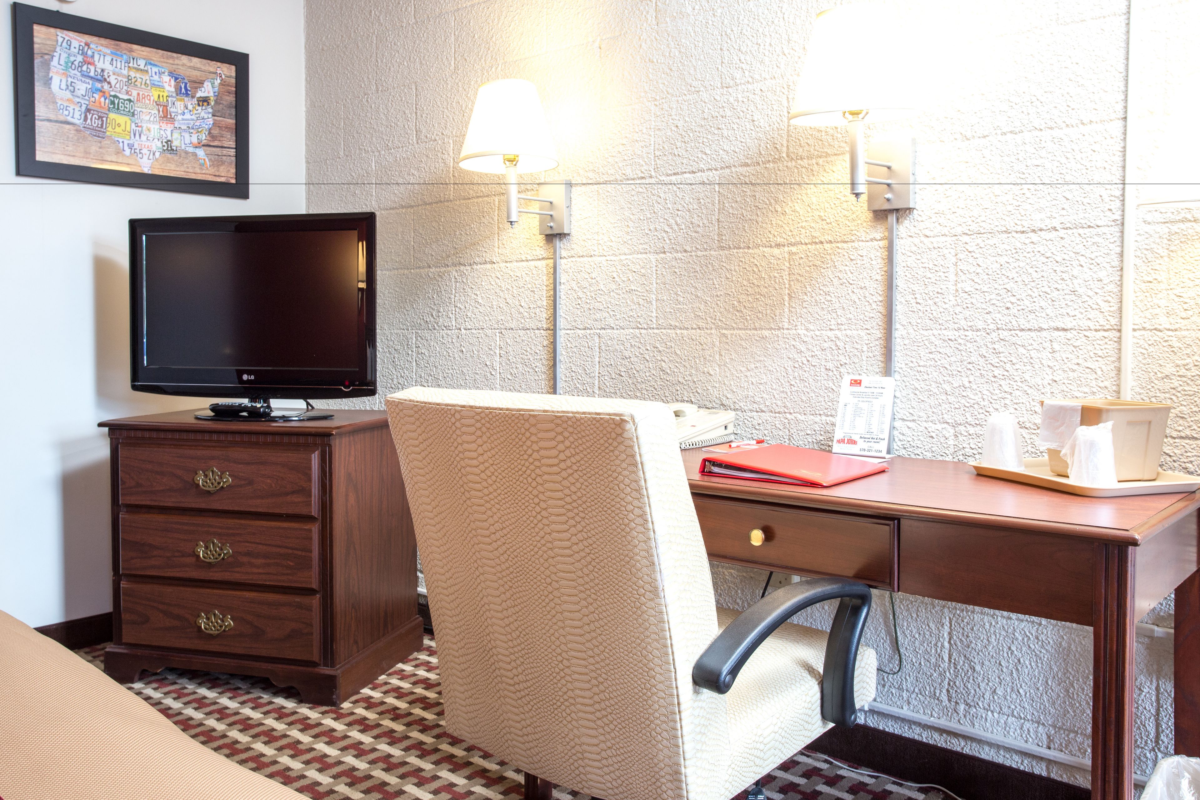 A hotel room with a wooden desk, a chair, a television, a lamp, and a framed map on the wall.