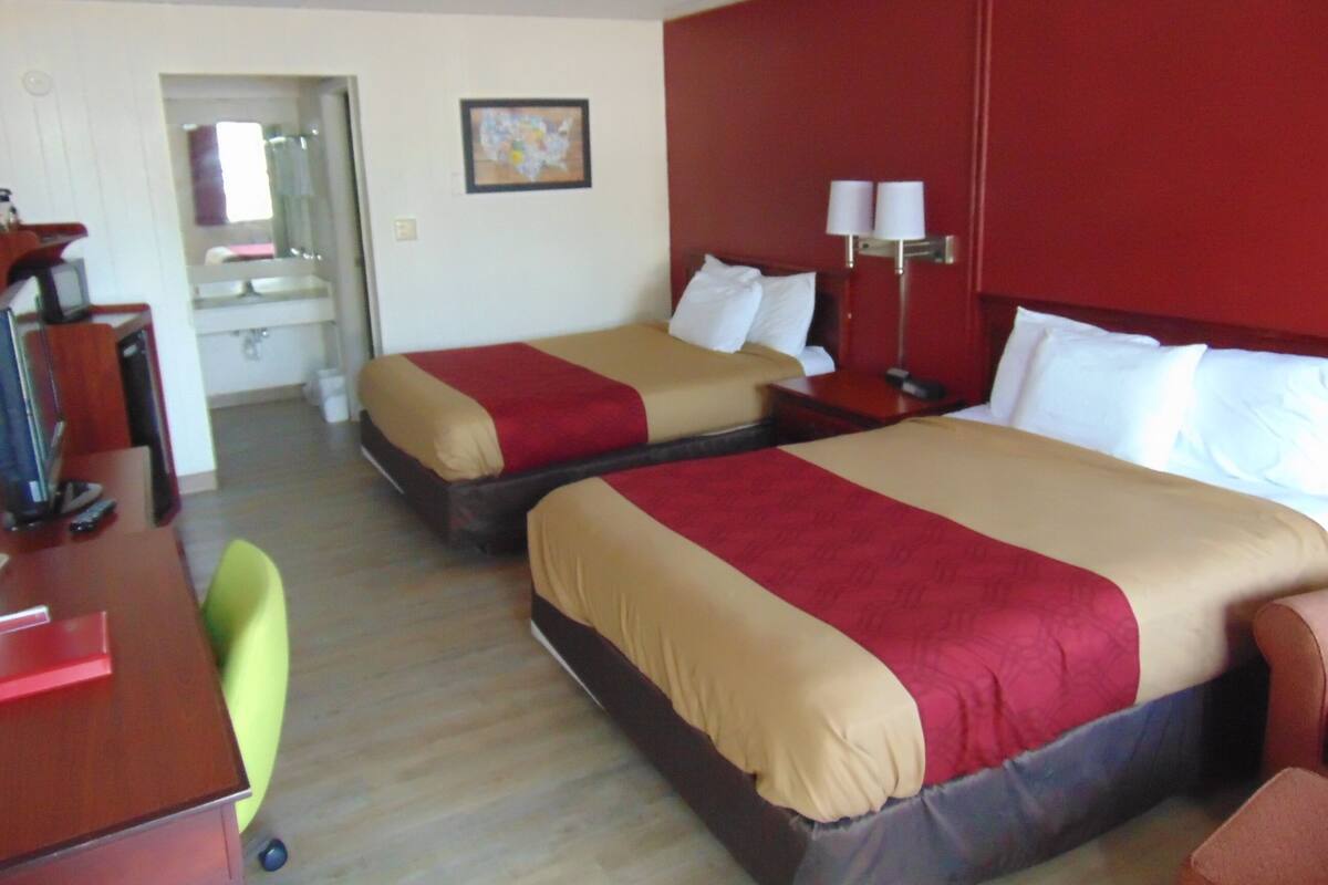 A hotel room with two beds, a desk, a chair, a lamp, and a bathroom visible through an open door.
