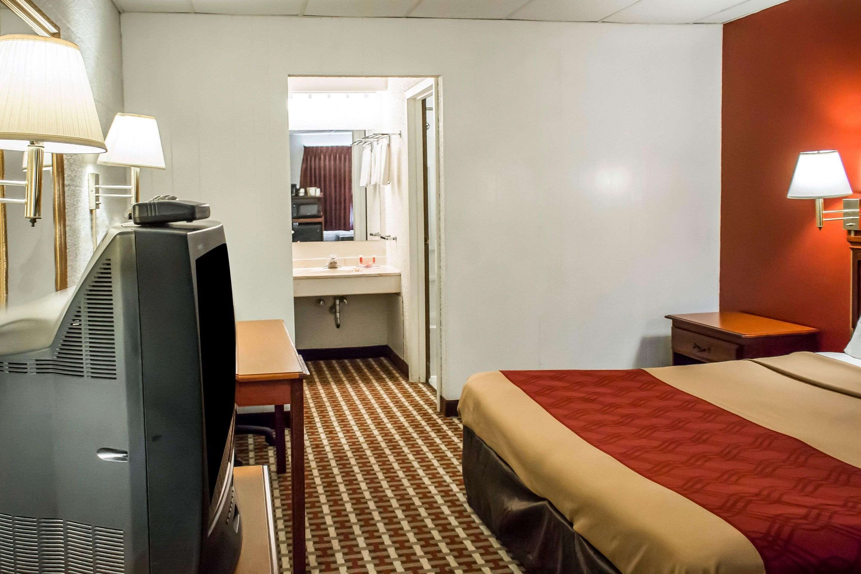 A hotel room with a bed, a desk, a television, a bathroom visible through an open door, and two wall-mounted lamps.