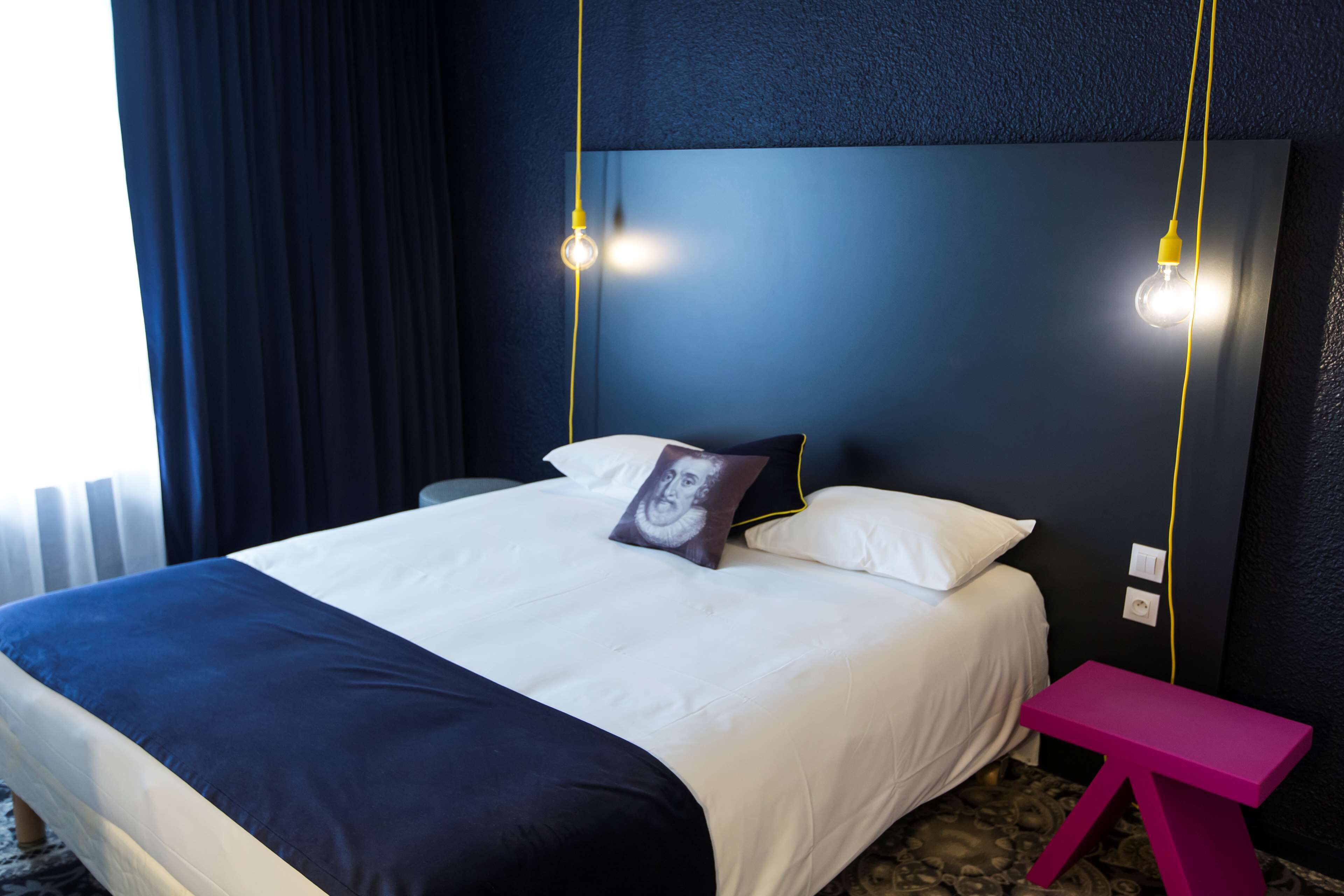 A hotel room with a bed, a nightstand, a pink bench, and two pendant lights.