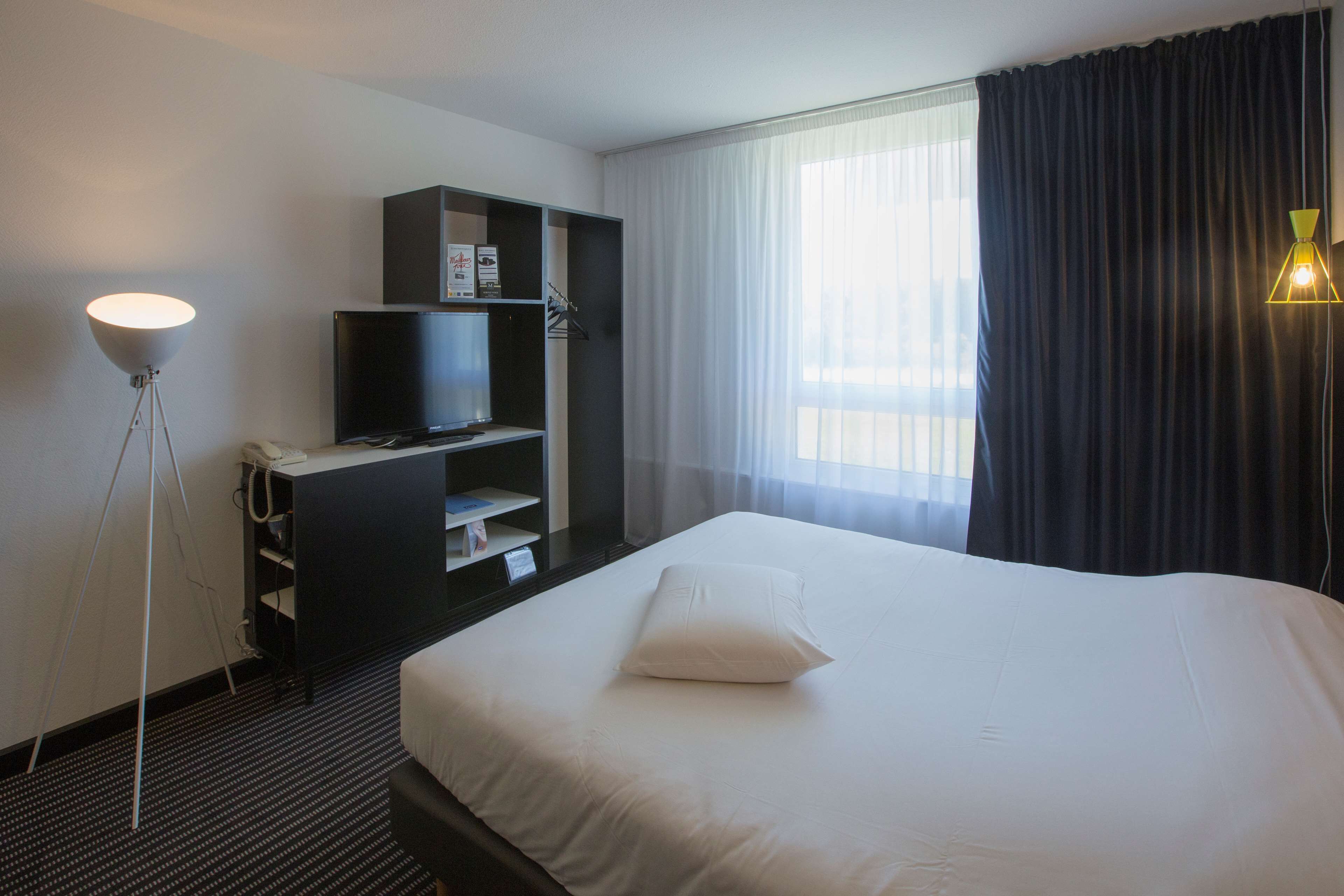 A hotel room with a large bed, a television on a stand, a desk with a chair, and a floor lamp.