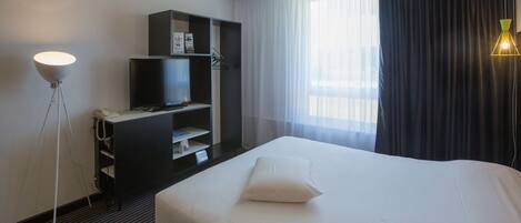 A hotel room with a large bed, a television on a stand, a desk with a chair, and a floor lamp.