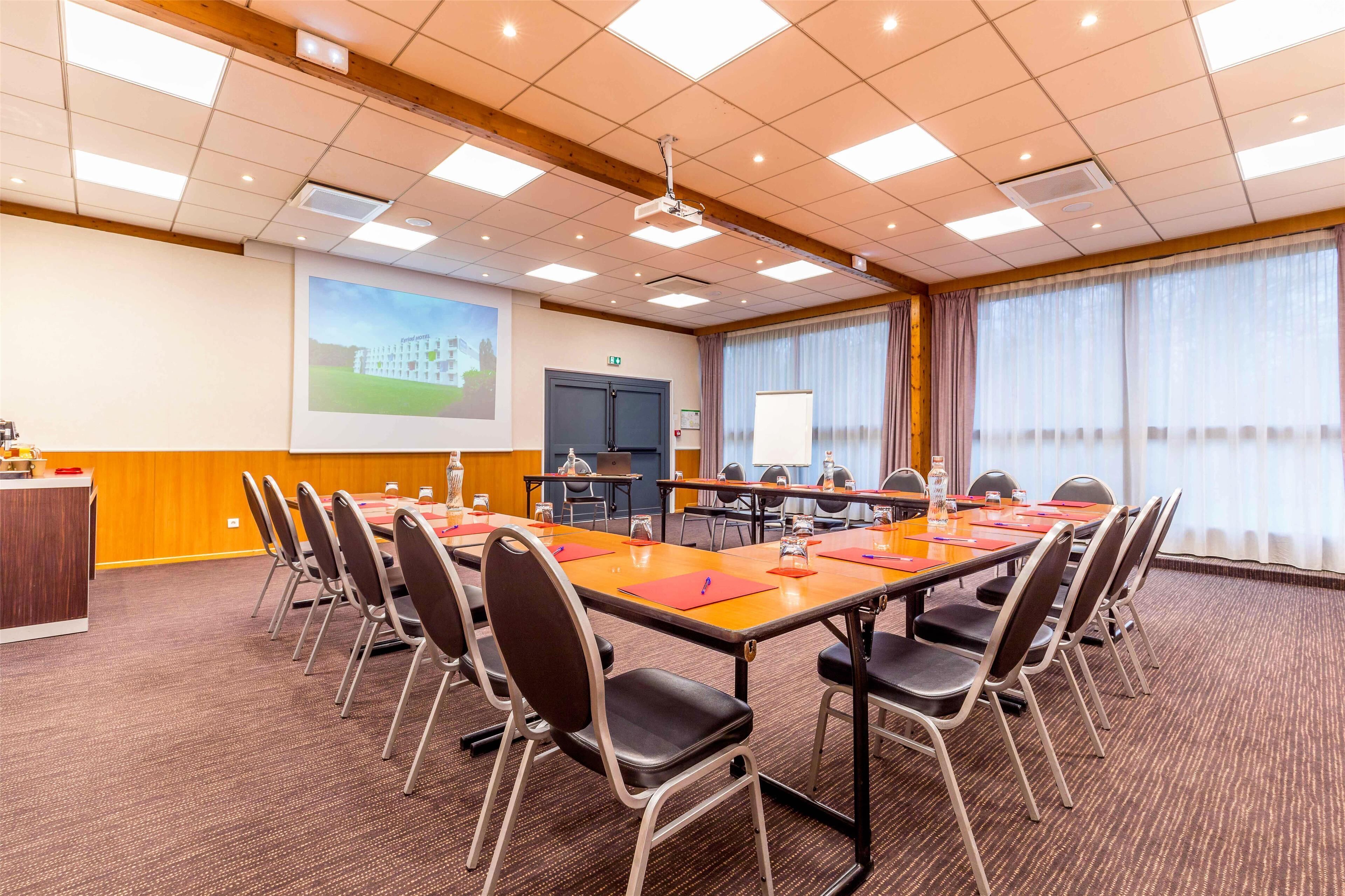 A conference room with a long table, chairs, a projector screen, and a large window with curtains.