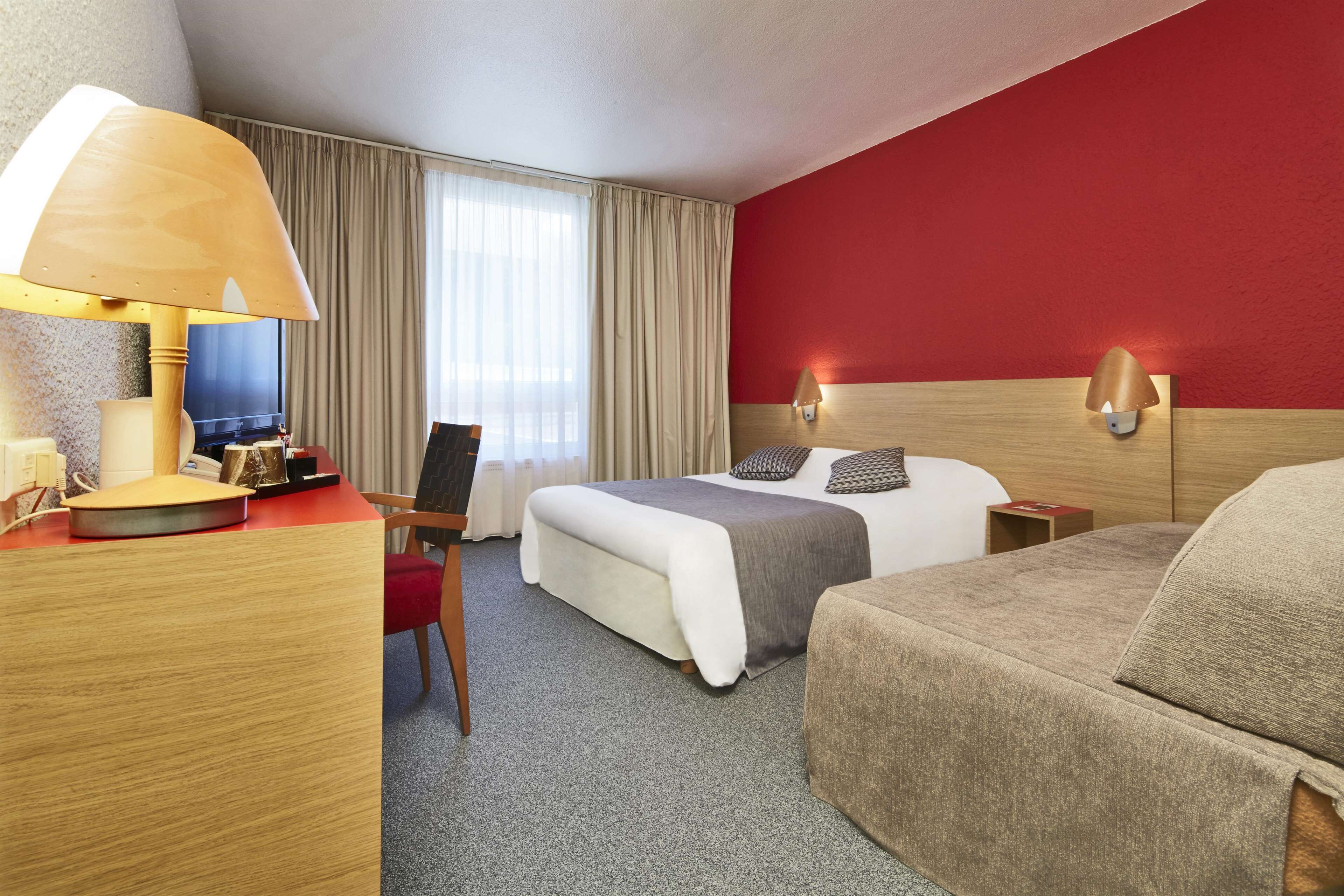 A hotel room with a bed, a desk with a lamp, a chair, a TV, and a red accent wall.