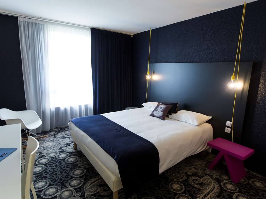 A hotel room with a bed, a desk, a chair, a small table, and a patterned carpet.