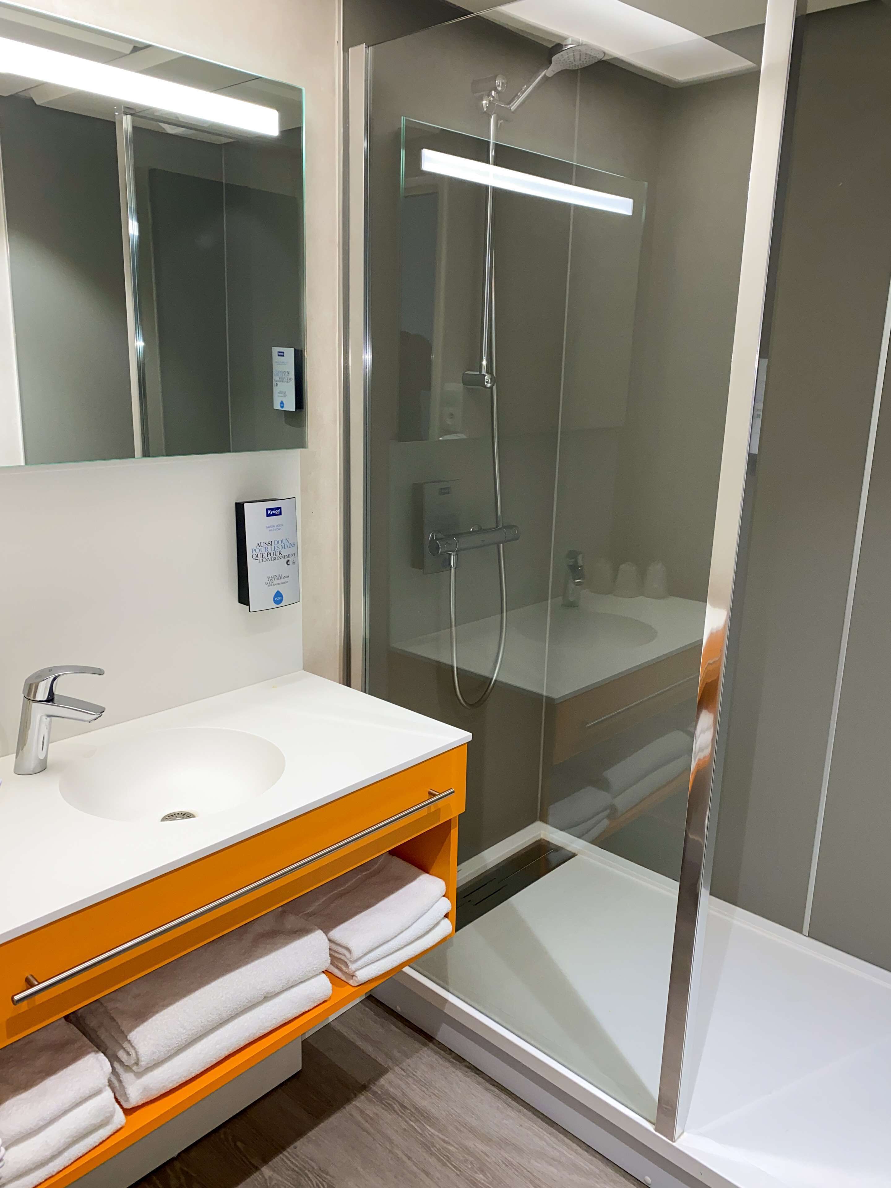 Une salle de bain moderne avec une douche à parois de verre, un lavabo blanc avec un meuble-lavabo orange, et un miroir au-dessus du lavabo.