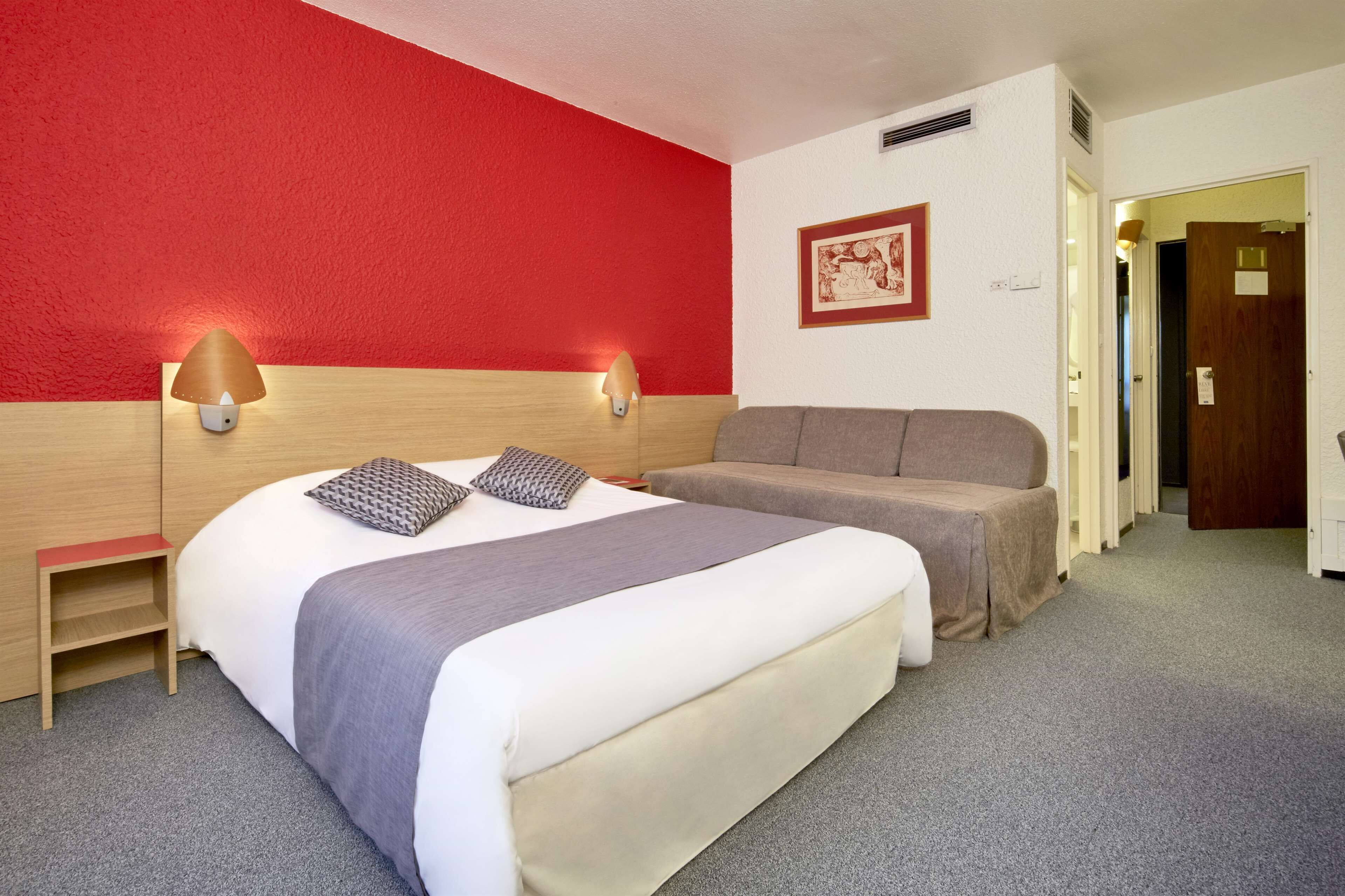 A hotel room with a bed, bedside tables, a sofa, and a red accent wall.