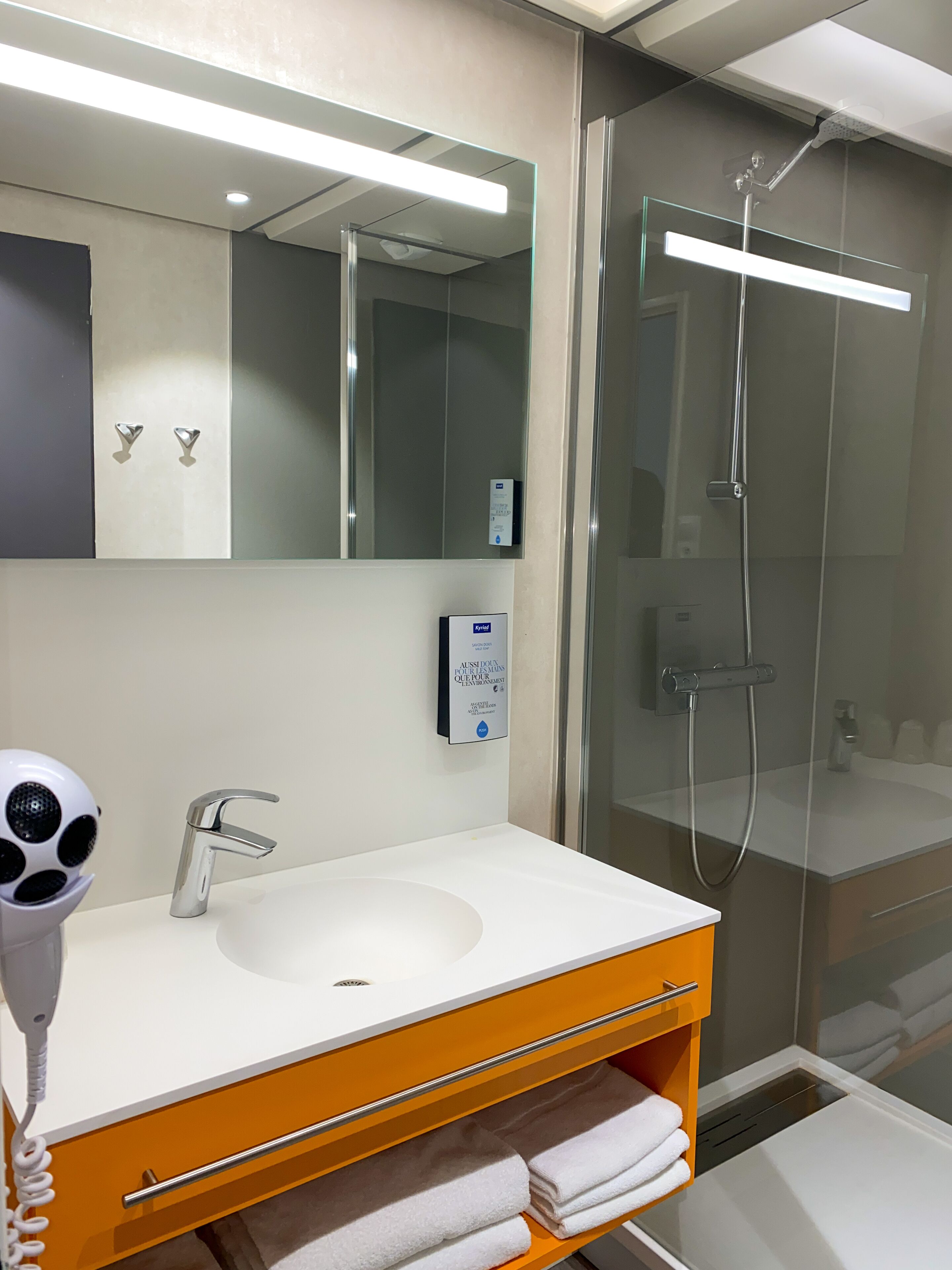 A modern bathroom with a white sink, a handheld hair dryer, a glass shower enclosure, and a mirror with a control panel.
