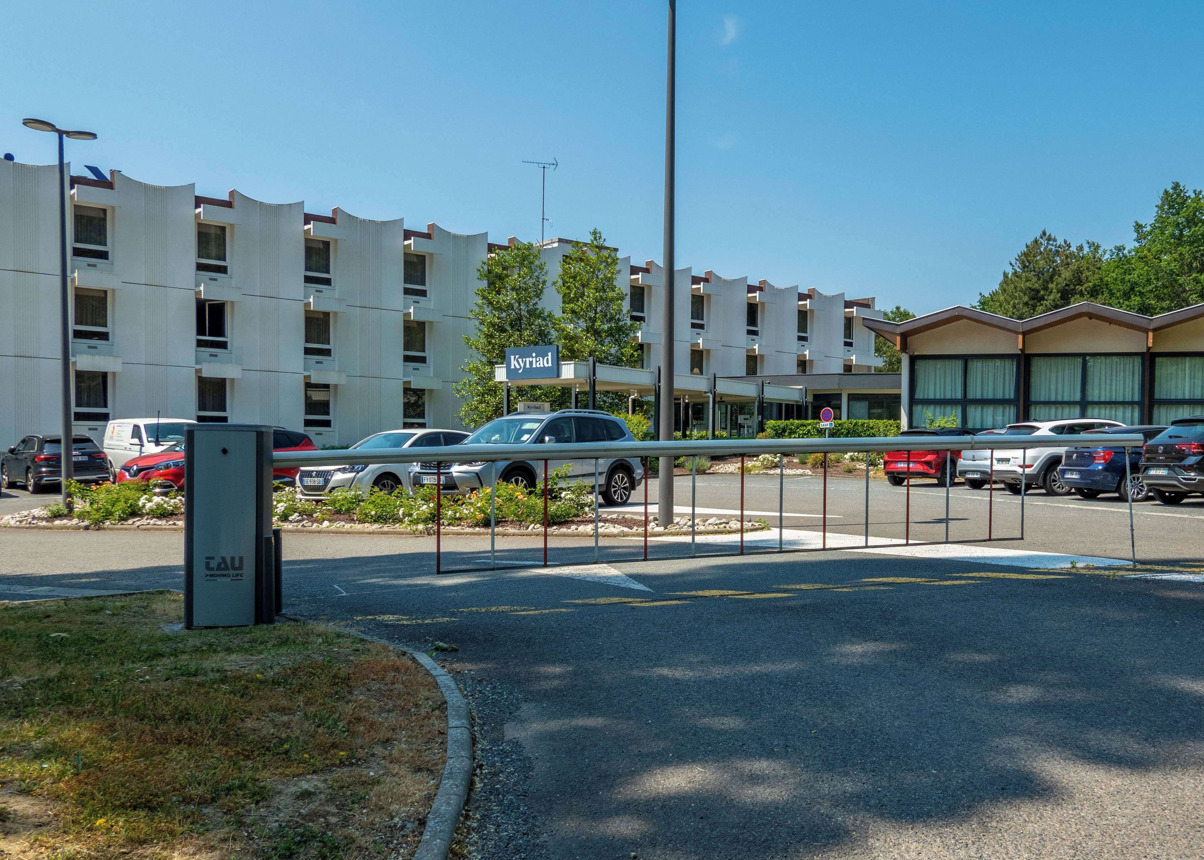 Un parking avec plusieurs voitures et, au fond, un bâtiment portant l’inscription « Kyriad ».