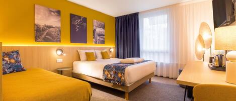 A hotel room with a bed, a desk, and two chairs. There are three wall-mounted artworks above the bed.