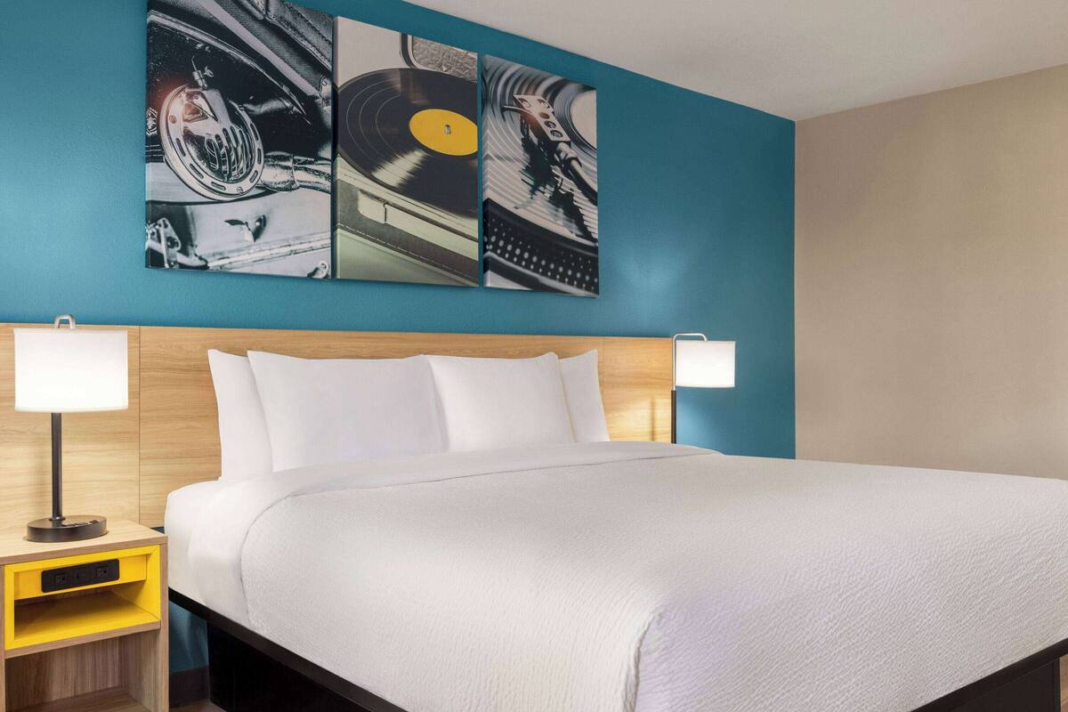 A hotel room with a large bed, two bedside lamps, a wooden headboard, and three wall-mounted artwork pieces.