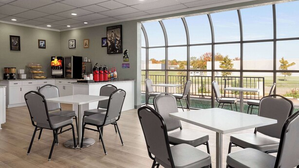 A modern dining area with white tables and chairs, a kitchen area with appliances, and large windows offering a view of the outdoors.