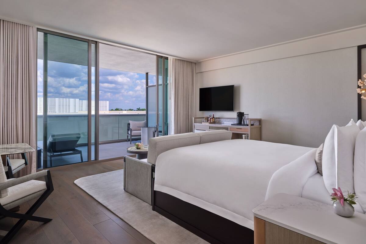 Ein modernes Hotelzimmer mit einem großen Bett, einem Schreibtisch, einem Sessel, einem Fernseher und einem Balkon mit Blick auf die Stadt.