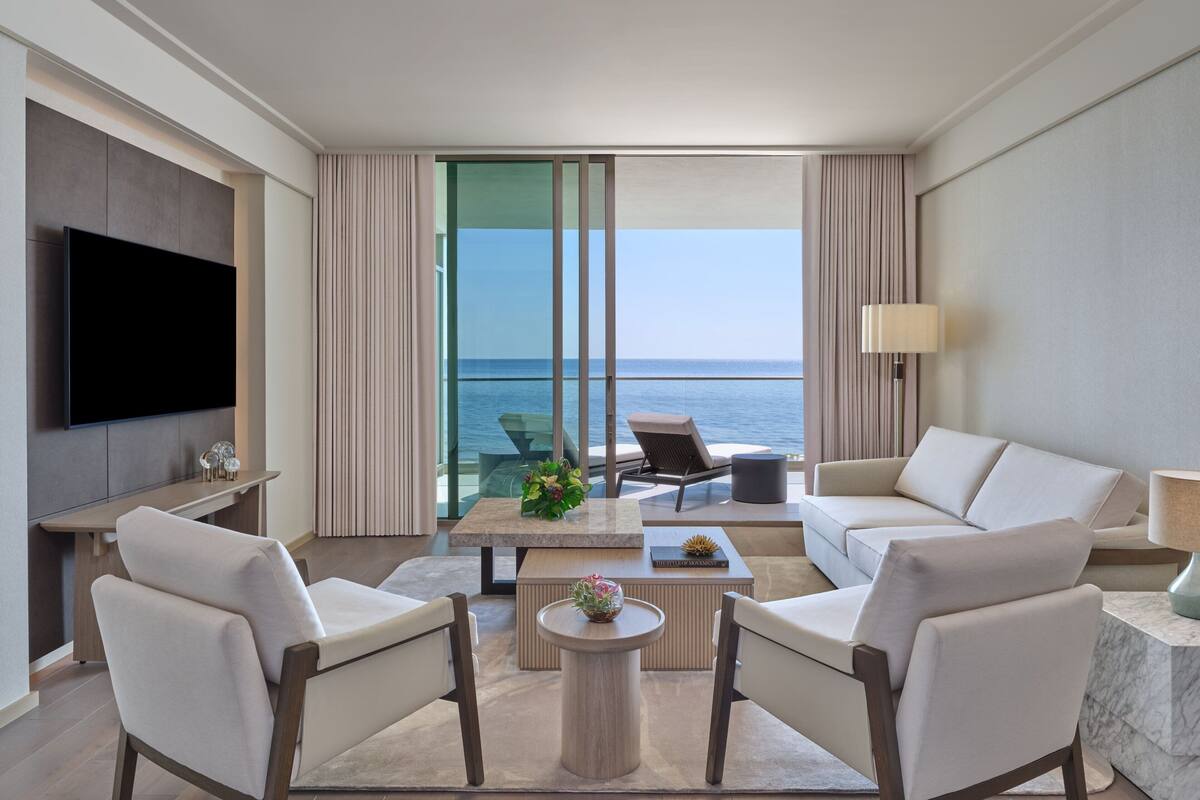 Ein modernes Hotelzimmer mit einem großen Flachbildfernseher, einer Couch, zwei Sesseln, einem Couchtisch und Meerblick durch raumhohe Fenster.