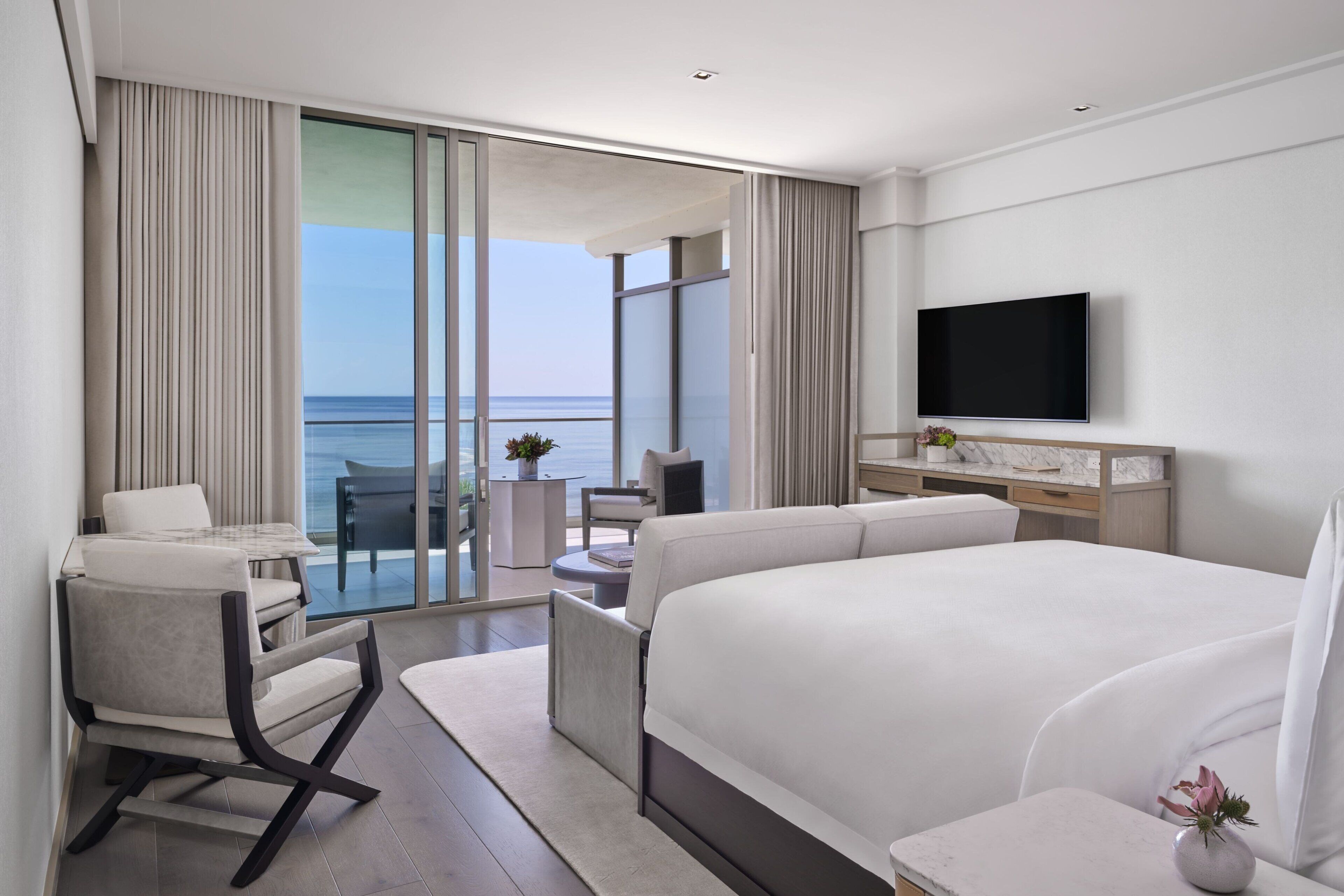 A modern hotel room with a large bed, a TV, a balcony with a view of the sea, and a seating area with a table and chairs.